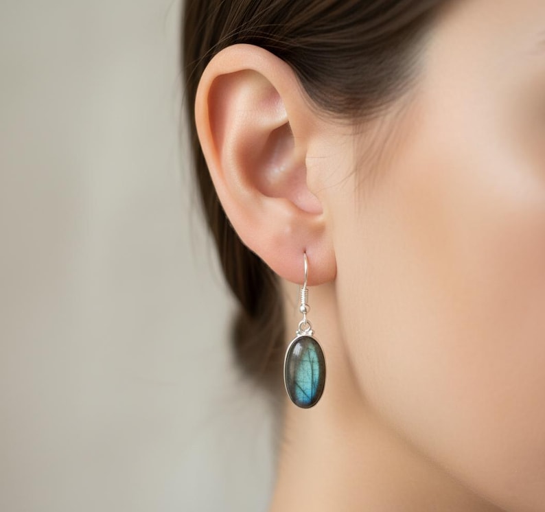 May include: A silver drop earring featuring an oval-shaped labradorite gemstone. The stone displays a dark, iridescent blue and grey surface with natural inclusions. The earring hangs from a simple silver hook wire, showcasing a polished finish.