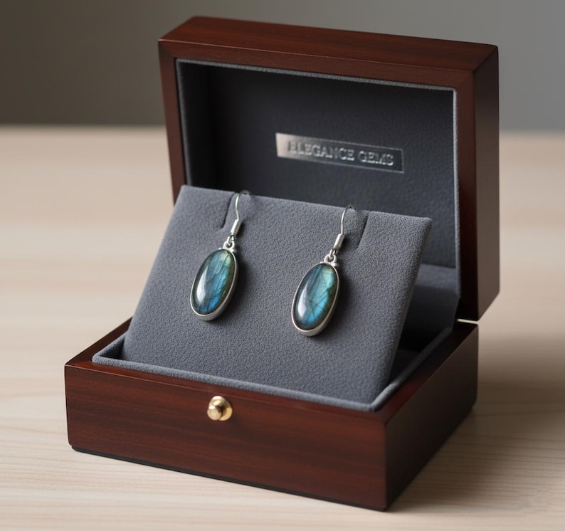 May include: A pair of oval-shaped labradorite drop earrings with silver hooks, displayed on a grey velvet insert inside a dark brown wooden jewelry box. The interior lid features a silver plaque with the text ELEGANCE GEMS in capital letters.