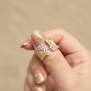 May include: A gold-toned ring with a textured finish, adorned with sparkling clear crystals. The ring is split into two sections, each embellished with the crystals. The design is elegant and eye-catching, perfect for special occasions.
