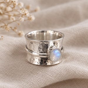 May include: A wide silver spinner ring featuring a hammered, textured finish on the main band. A smooth, polished silver band rotates around the centre, accented by a single, round, cabochon-cut moonstone that displays a soft blue iridescence.