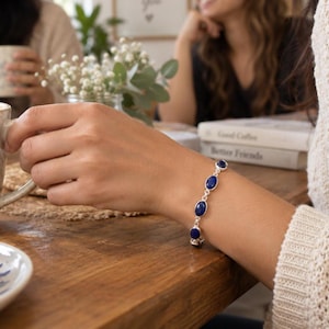 May include: A silver chain bracelet featuring five oval-shaped, deep blue lapis lazuli gemstones set in polished silver bezels. The bracelet is secured with a silver link chain, showcasing a classic design that highlights the vibrant blue stones.