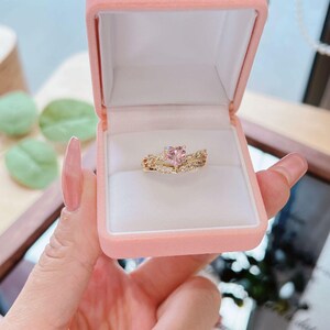 May include: A gold ring set with a pink heart-shaped gemstone, displayed in a pink velvet ring box. The ring is surrounded by smaller clear gemstones. The box is held open, showcasing the jewelry.