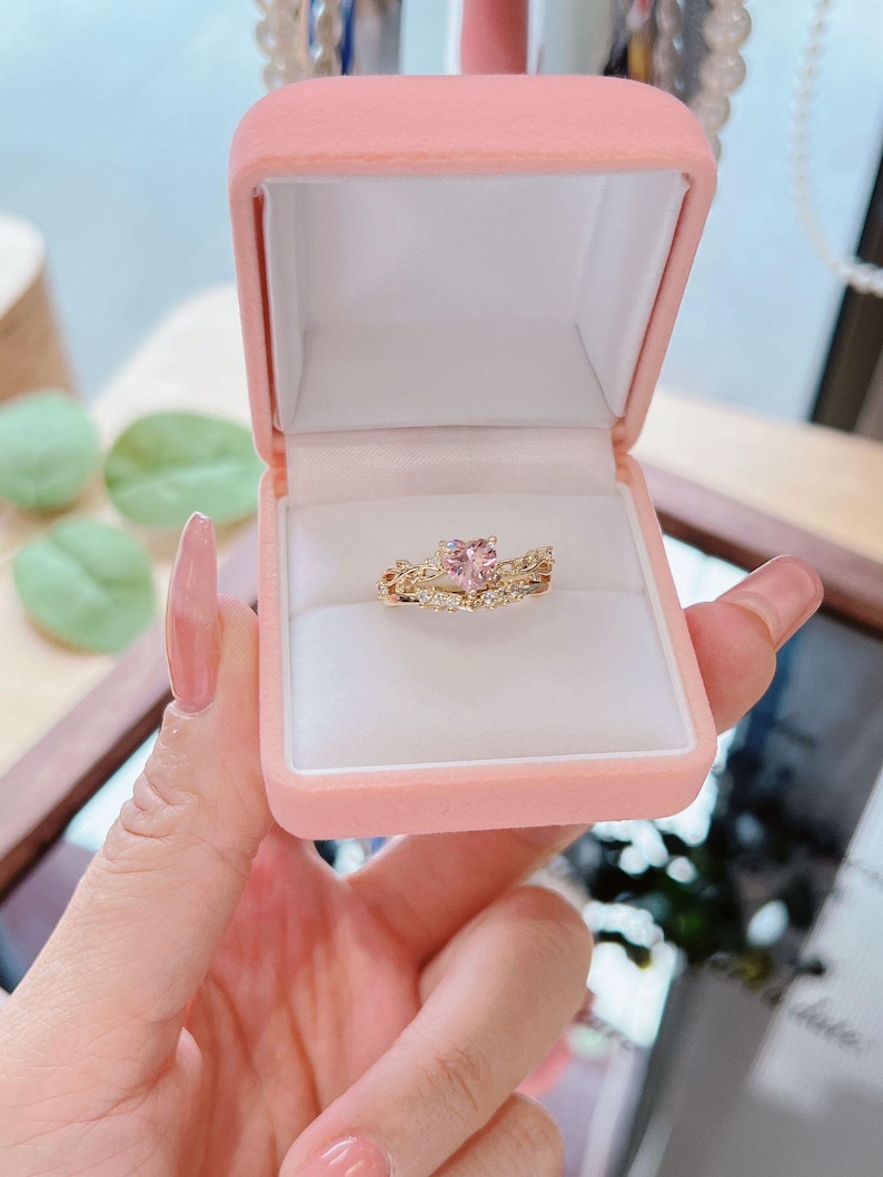 May include: A gold ring set with a pink heart-shaped gemstone, displayed in a pink velvet ring box. The ring is surrounded by smaller clear gemstones. The box is held open, showcasing the jewelry.
