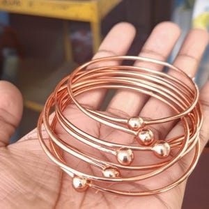 Op de afbeelding: Een verzameling koperkleurige armbanden. Elke armband heeft een glad, afgerond ontwerp met een klein, bolvormig accent. De armbanden zijn op elkaar gestapeld en tonen hun eenvoudige maar elegante stijl. De warme, metallic tint suggereert een klassiek sieraad.