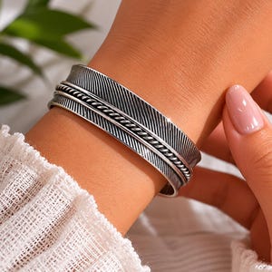 May include: A silver-toned cuff bracelet with a feather design. The bracelet features detailed feather etching and a braided rope-like accent. The cuff is approximately 1.5 inches wide and is worn on a wrist with a white sweater.