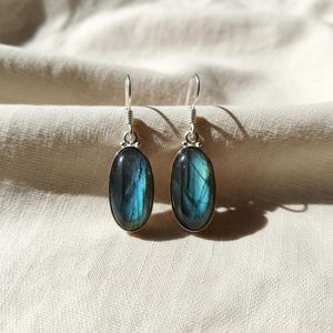 May include: A pair of drop earrings featuring oval-shaped labradorite gemstones with blue and grey iridescence. Each stone is set in a polished silver-toned metal bezel with a simple hook wire attachment, showcasing natural stone variations.