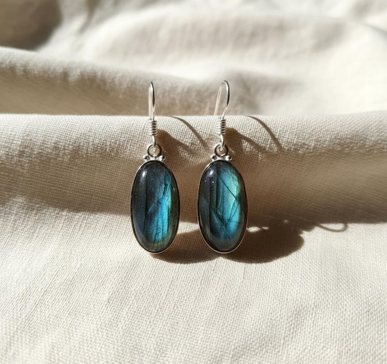 May include: A pair of drop earrings featuring oval-shaped labradorite gemstones with blue and grey iridescence. Each stone is set in a polished silver-toned metal bezel with a simple hook wire attachment, showcasing natural stone variations.