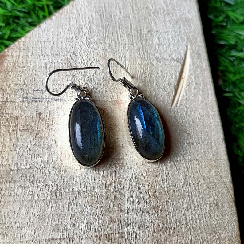 May include: A pair of silver drop earrings featuring oval-shaped labradorite gemstones. Each earring has a polished, dark grey stone with iridescent blue flashes set in a simple silver bezel, suspended from a curved silver ear wire hook.