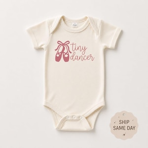 May include: A cream-colored short-sleeve baby bodysuit featuring a graphic of pink ballet slippers and the text "tiny dancer" printed in a matching pink script font on the chest. The garment has a round neckline and snap closures at the bottom.