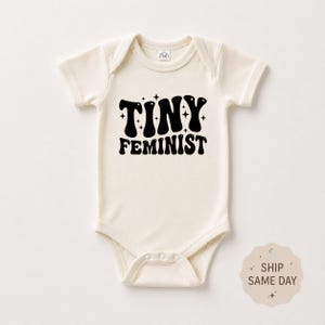 May include: A cream-colored short-sleeve baby bodysuit featuring the black text "TINY FEMINIST" in a bold, wavy font, accented by small black star icons. The garment has a round neckline and snap closures at the bottom for easy access.
