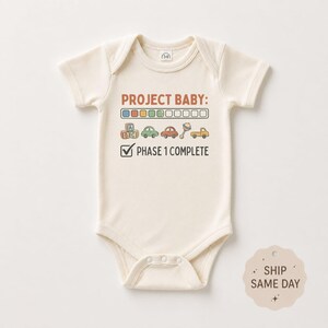 May include: A cream-colored short-sleeve baby bodysuit featuring the text PROJECT BABY: followed by a progress bar with four colored squares, illustrations of building blocks, cars, and a rattle, and the text PHASE 1 COMPLETE with a checkmark.