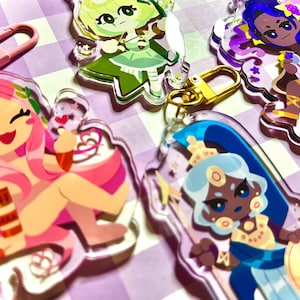 May include: A collection of colorful acrylic keychains featuring cartoon characters. The keychains have gold and pink clasps. The characters have pink, green, blue, and purple hair and outfits. The keychains are on a purple and white checkered surface.