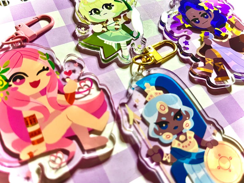 May include: A collection of colorful acrylic keychains featuring cartoon characters. The keychains have gold and pink clasps. The characters have pink, green, blue, and purple hair and outfits. The keychains are on a purple and white checkered surface.