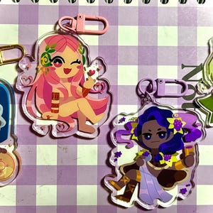 May include: A collection of colorful acrylic keychains featuring cartoon illustrations of female figures. Each keychain has a different design, including a figure with pink hair, a figure with blue hair, and a figure with green hair. Each keychain has a metal clasp.