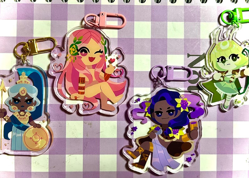 May include: A collection of colorful acrylic keychains featuring cartoon illustrations of female figures. Each keychain has a different design, including a figure with pink hair, a figure with blue hair, and a figure with green hair. Each keychain has a metal clasp.