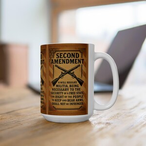 Pode incluir: Caneca de cerâmica branca com o texto da Segunda Emenda e um desenho de rifles cruzados. A caneca tem um fundo castanho e bege com o texto "Uma milícia bem regulamentada, sendo necessária para a segurança de um Estado livre. O direito do povo de ter e portar armas não será infringido."