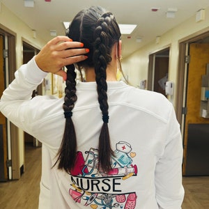 May include: White long-sleeved shirt with a graphic print of medical supplies and the word "NURSE" on the back. The person is wearing their hair in two braids.