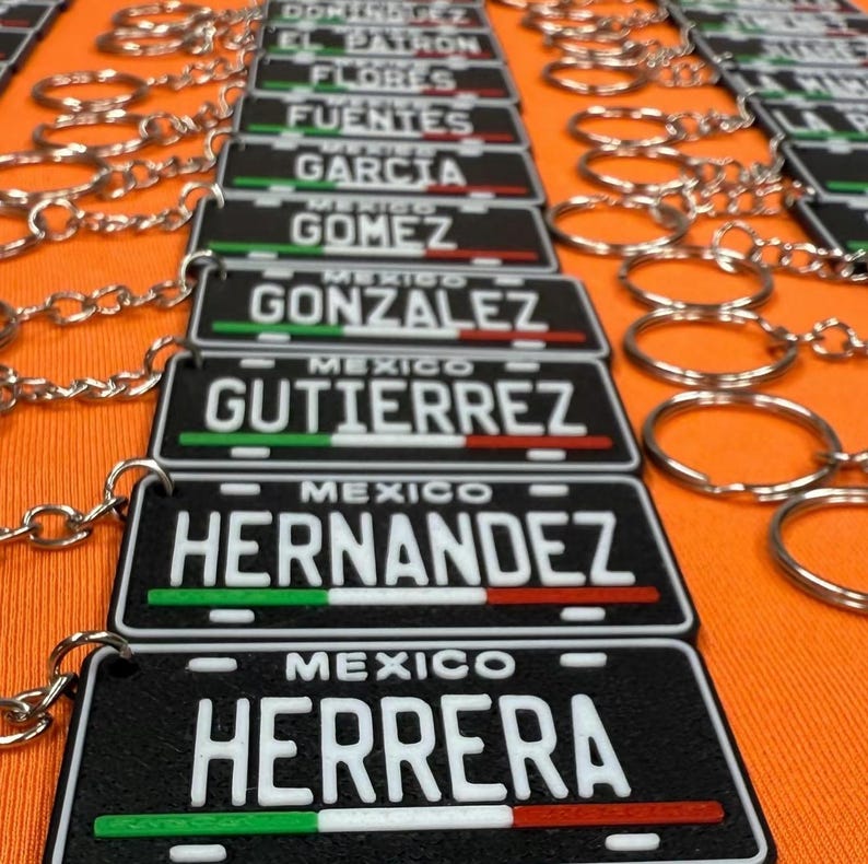 K&ouml;nnte beinhalten: Schwarze Schl&uuml;sselanh&auml;nger mit wei&szlig;en Familiennamen und dem Wort "MEXICO" dar&uuml;ber. Die Schl&uuml;sselanh&auml;nger haben einen gr&uuml;nen, wei&szlig;en und roten Streifen. Sie sind an silbernen Schl&uuml;sselringen befestigt und liegen auf einer orangefarbenen Oberfl&auml;che.