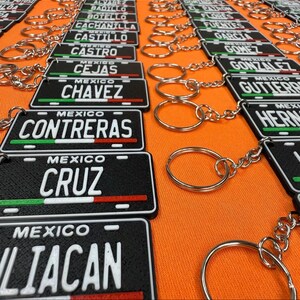 K&ouml;nnte beinhalten: Schwarze Schl&uuml;sselanh&auml;nger mit rechteckigem Kennzeichen-Design. Jeder Schl&uuml;sselanh&auml;nger zeigt einen Namen in wei&szlig;en Buchstaben, das Wort "MEXICO" dar&uuml;ber und einen gr&uuml;nen, wei&szlig;en und roten Streifen unten. Die Schl&uuml;sselanh&auml;nger liegen auf einer orangefarbenen Oberfl&auml;che.