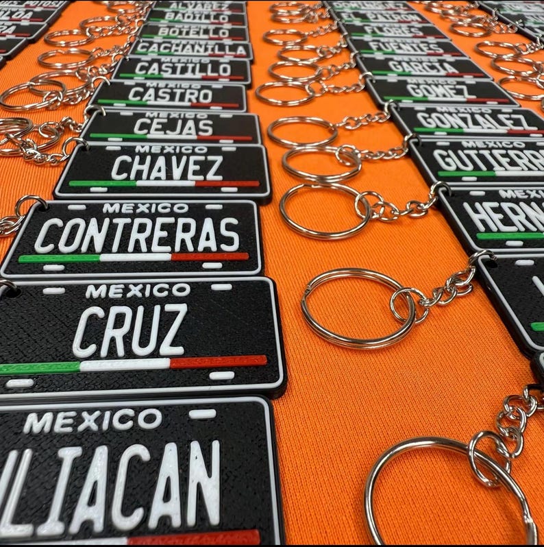 K&ouml;nnte beinhalten: Schwarze Schl&uuml;sselanh&auml;nger mit rechteckigem Kennzeichen-Design. Jeder Schl&uuml;sselanh&auml;nger zeigt einen Namen in wei&szlig;en Buchstaben, das Wort "MEXICO" dar&uuml;ber und einen gr&uuml;nen, wei&szlig;en und roten Streifen unten. Die Schl&uuml;sselanh&auml;nger liegen auf einer orangefarbenen Oberfl&auml;che.
