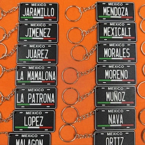 K&ouml;nnte beinhalten: Schwarze Schl&uuml;sselanh&auml;nger mit kleinen Nummernschildern mit Namen wie JARAMILLO und JIMENEZ. Jedes Schild tr&auml;gt die Aufschrift "MEXICO" und ein gr&uuml;nes, wei&szlig;es und rotes Streifendesign. Der Hintergrund ist orange.