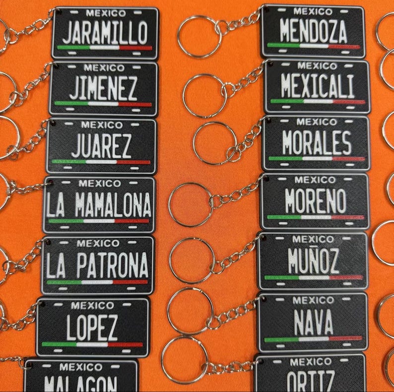 K&ouml;nnte beinhalten: Schwarze Schl&uuml;sselanh&auml;nger mit kleinen Nummernschildern mit Namen wie JARAMILLO und JIMENEZ. Jedes Schild tr&auml;gt die Aufschrift "MEXICO" und ein gr&uuml;nes, wei&szlig;es und rotes Streifendesign. Der Hintergrund ist orange.