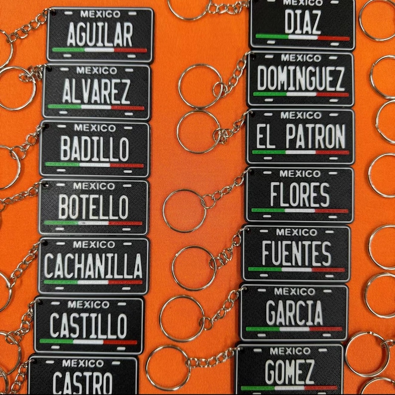 K&ouml;nnte beinhalten: Schwarze Schl&uuml;sselanh&auml;nger mit Miniatur-Kennzeichen mit Namen und dem Wort "MEXICO". Jedes Schild hat ein Design mit gr&uuml;nen, wei&szlig;en und roten Streifen, mit Namen wie AGUILAR, DIAZ und DOMINGUEZ. Der Hintergrund ist orange.