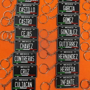 K&ouml;nnte beinhalten: Eine Sammlung schwarzer Schl&uuml;sselanh&auml;nger im Nummernschild-Design. Jeder Schl&uuml;sselanh&auml;nger zeigt einen Namen und das Wort "MEXICO" &uuml;ber einem gr&uuml;nen, wei&szlig;en und roten Streifen. Die Schl&uuml;sselanh&auml;nger sind mit einer silbernen Kette und einem Ring verbunden.