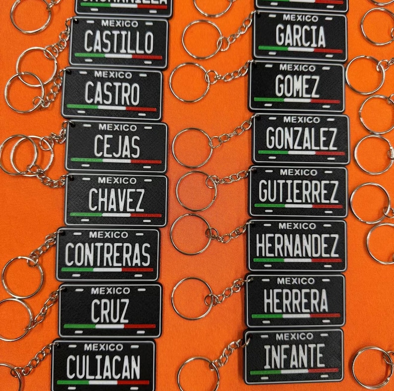K&ouml;nnte beinhalten: Eine Sammlung schwarzer Schl&uuml;sselanh&auml;nger im Nummernschild-Design. Jeder Schl&uuml;sselanh&auml;nger zeigt einen Namen und das Wort "MEXICO" &uuml;ber einem gr&uuml;nen, wei&szlig;en und roten Streifen. Die Schl&uuml;sselanh&auml;nger sind mit einer silbernen Kette und einem Ring verbunden.