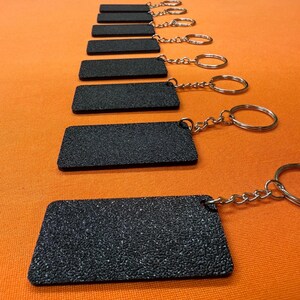 K&ouml;nnte beinhalten: Eine Reihe schwarzer, rechteckiger Schl&uuml;sselanh&auml;nger mit strukturierter Oberfl&auml;che, jeweils mit einer silbernen Kette und einem Ring verbunden. Die Schl&uuml;sselanh&auml;nger sind auf orangefarbenem Hintergrund angeordnet.