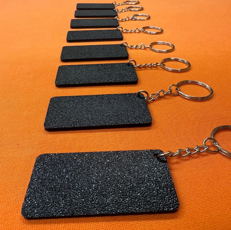K&ouml;nnte beinhalten: Eine Reihe schwarzer, rechteckiger Schl&uuml;sselanh&auml;nger mit strukturierter Oberfl&auml;che, jeweils mit einer silbernen Kette und einem Ring verbunden. Die Schl&uuml;sselanh&auml;nger sind auf orangefarbenem Hintergrund angeordnet.