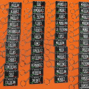 K&ouml;nnte beinhalten: Eine Sammlung von Miniatur-Kennzeichen-Schl&uuml;sselanh&auml;ngern. Jedes schwarze Schild zeigt einen Namen und das Wort "MEXICO" &uuml;ber einem gr&uuml;nen, wei&szlig;en und roten Streifen. Die Schl&uuml;sselanh&auml;nger sind an silbernen Schl&uuml;sselringen befestigt.