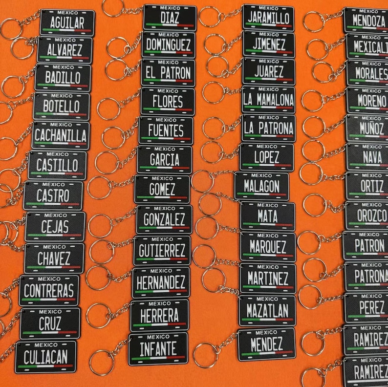 K&ouml;nnte beinhalten: Eine Sammlung von Miniatur-Kennzeichen-Schl&uuml;sselanh&auml;ngern. Jedes schwarze Schild zeigt einen Namen und das Wort "MEXICO" &uuml;ber einem gr&uuml;nen, wei&szlig;en und roten Streifen. Die Schl&uuml;sselanh&auml;nger sind an silbernen Schl&uuml;sselringen befestigt.