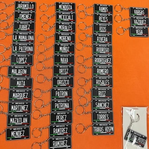 K&ouml;nnte beinhalten: Eine Sammlung schwarzer Schl&uuml;sselanh&auml;nger mit wei&szlig;en Schriftz&uuml;gen, die kleinen Nummernschildern &auml;hneln. Jeder Schl&uuml;sselanh&auml;nger zeigt einen anderen Nachnamen mit dem Wort "MEXICO" dar&uuml;ber. Die Schl&uuml;sselanh&auml;nger sind an silbernen Schl&uuml;sselringen befestigt.