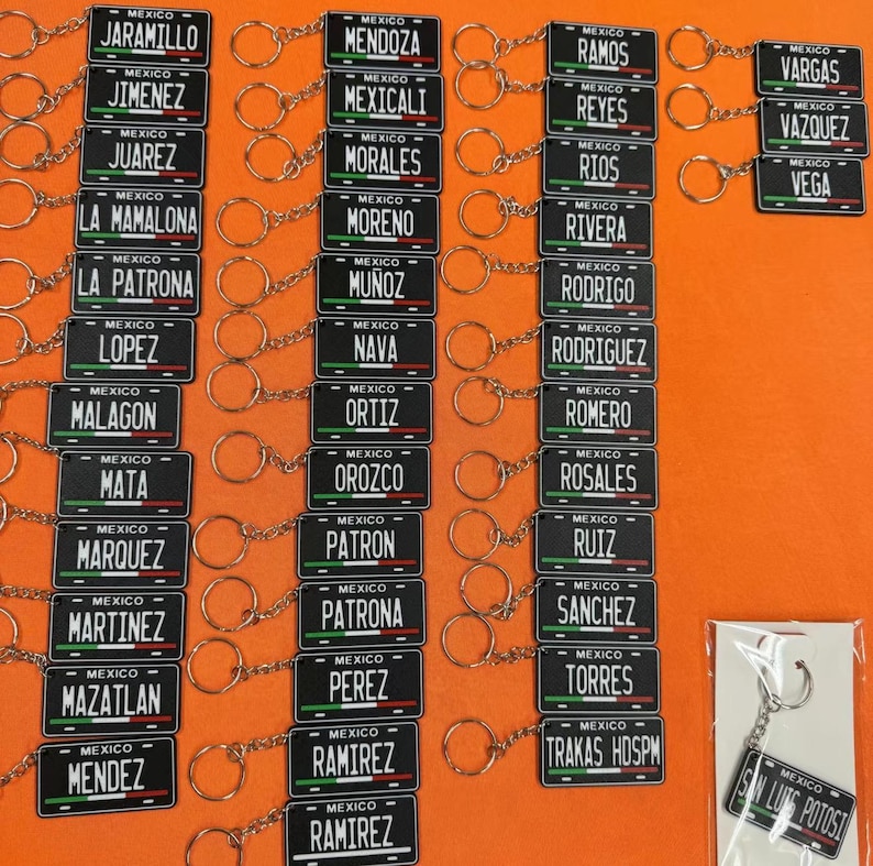 K&ouml;nnte beinhalten: Eine Sammlung schwarzer Schl&uuml;sselanh&auml;nger mit wei&szlig;en Schriftz&uuml;gen, die kleinen Nummernschildern &auml;hneln. Jeder Schl&uuml;sselanh&auml;nger zeigt einen anderen Nachnamen mit dem Wort "MEXICO" dar&uuml;ber. Die Schl&uuml;sselanh&auml;nger sind an silbernen Schl&uuml;sselringen befestigt.