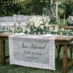 Puede incluir: Una mesa rústica de madera preparada para una recepción de boda. La mesa está decorada con flores blancas, velas y un camino de mesa transparente con el texto "Just Married", los nombres "Emily & Jake" y el año "est. 2025".