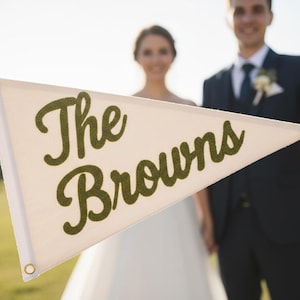 Puede incluir: Bandera triangular crema con las palabras "The Browns" en escritura verde. La bandera está sostenida por dos personas, con una novia y un novio en el fondo. Decoración de boda.