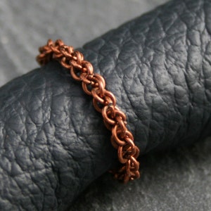 Könnte beinhalten: Ein Kupferkettenring mit geflochtenem Design. Der Ring liegt auf einer dunkelgrauen, strukturierten Oberfläche.