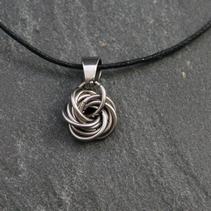 Könnte beinhalten: Eine silberne Halskette mit einem schwarzen Band und einem silbernen Anhänger in Form eines Knotens.