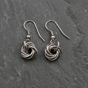 Könnte beinhalten: Ein Paar silberne Ohrringe mit einem gedrehten Knotendesign. Die Ohrringe sind aus Metall und haben einen schlichten, eleganten Stil.