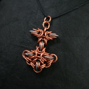 May include: A copper and silver chainmail pendant in the shape of a stylized hammer, hanging from a black cord.
