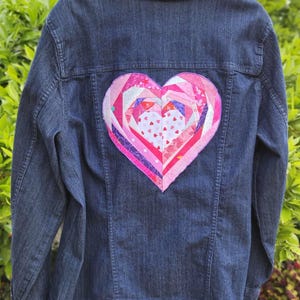 Puede incluir: Una chaqueta vaquera azul oscuro con un gran parche en forma de corazón rosa y blanco cosido en la espalda. El corazón está hecho de múltiples piezas de tela cosidas juntas en espiral.