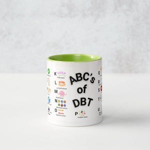 May include: White ceramic mug with a green interior, featuring the text "ABC's of DBT" in black letters. The mug is decorated with colourful illustrations and text related to DBT concepts. The mug is on a white surface.