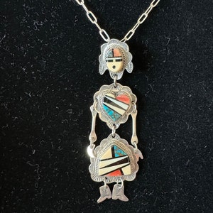 May include: A silver necklace with a pendant featuring three geometric shapes inlaid with turquoise, coral, and black stones. The pendant is in the shape of a face, a heart, and a stylized figure.