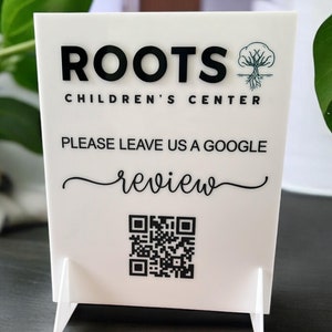 May include: White sign with black text that reads "ROOTS CHILDREN'S CENTER" and "PLEASE LEAVE US A GOOGLE review". A green tree with roots logo is in the top right corner. A QR code is below the text.