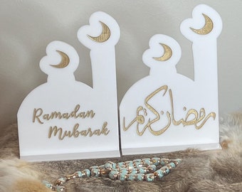 May include: Two white wooden mosque-shaped decorations with gold crescent moons and Arabic calligraphy. The decoration on the left says "Ramadan Mubarak" and the decoration on the right says "Ramadan Kareem".