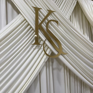 May include: A gold-coloured, reflective acrylic monogram featuring the letters K and S intertwined, mounted on a backdrop of white fabric drapes with vertical pleats. The letters are positioned centrally, creating a layered, decorative display.
