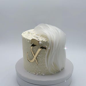 Peut inclure: Un gâteau blanc avec une lettre dorée 'X' sur le dessus. Le gâteau est décoré de perles blanches et a une frange de tissu blanc sur le dessus.