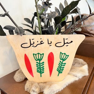 Puede incluir: Un macetero de madera con forma de taza de té marroquí tradicional con el texto árabe "شكل يا غزال" que significa "Hermosa, oh gacela" en letras negras. El macetero está decorado con tres flores estilizadas verdes y hojas rojas.