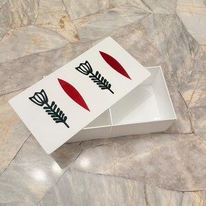 May include: A white rectangular box with a lid featuring a design of black floral illustrations and red leaf shapes. The box is open, revealing a white interior with two compartments. The box is on a marble surface.